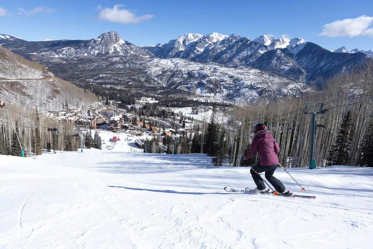 A skier descends a snow-covered mountain slope, with the majestic peaks of the surrounding range in the background. Below, a picturesque ski resort village nestles in the valley, capturing the spirit of mountain adventure and natural beauty.