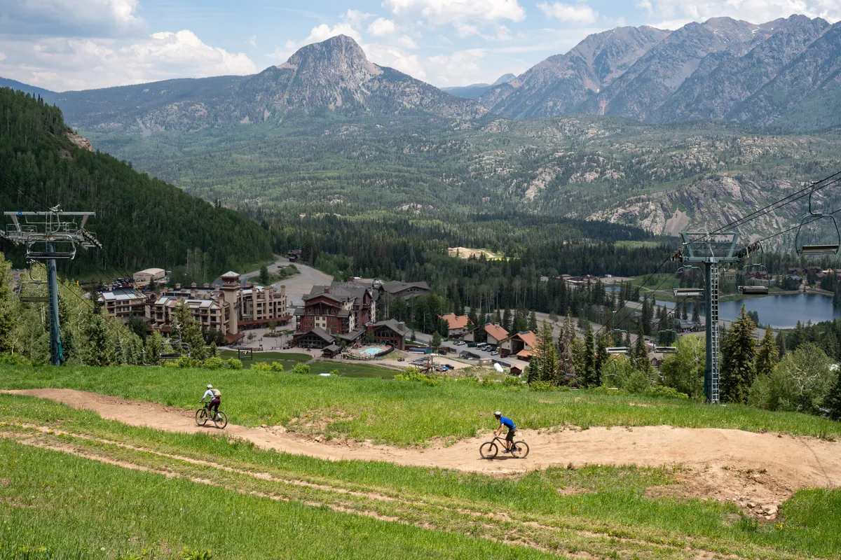 A sweeping exterior view captures dramatic mountain peaks, a biking trail, resort buildings, a tranquil lake, and a chairlift. The image showcases the grandeur of the Needles Mountains and San Juans, with recreational opportunities visible in the landscape.