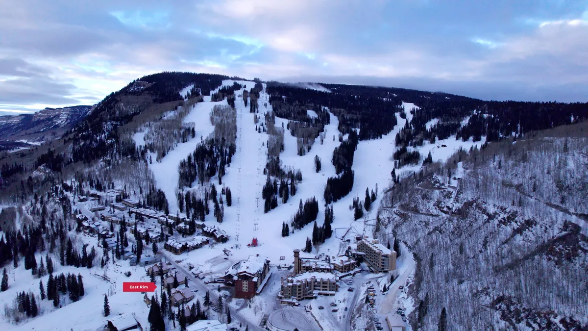 A panoramic view of snow-covered slopes with ski lifts ascending the mountain, resort buildings nestled at the base, and a dramatic mountain backdrop. The scene is alive with activity, as skiers and snowboarders take advantage of the fresh powder while sunlight glimmers off the snow.