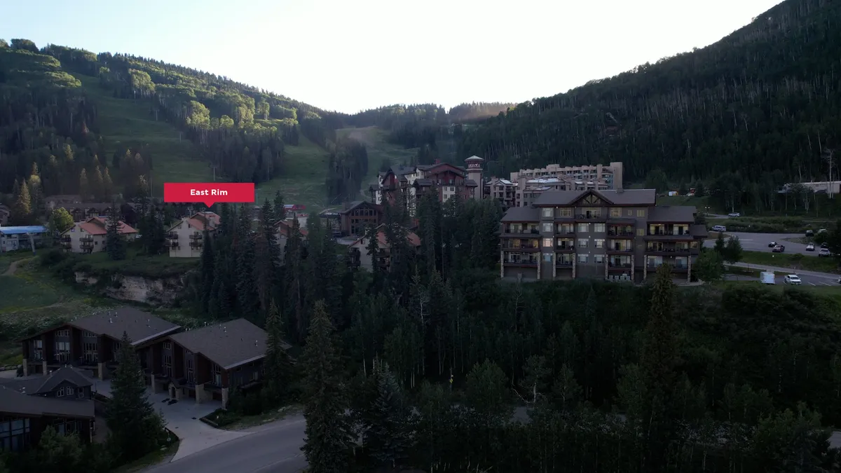 A panoramic exterior shot showing lush green hills, modern resort buildings, dense surrounding forests, and a roadway winding through the landscape. The scene exudes tranquility and hints at accessible hiking and exploration opportunities in the San Juans.
