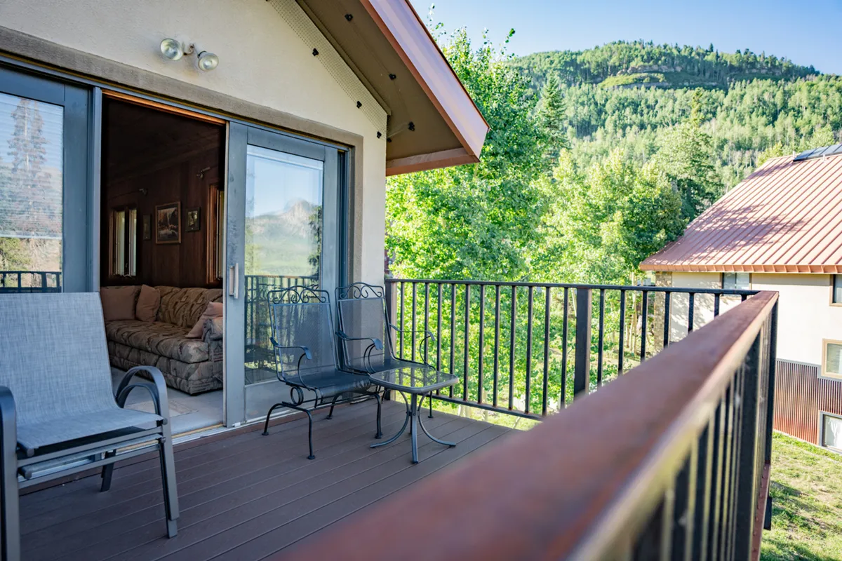 This image features a balcony seating area with comfortable chairs, set against a breathtaking backdrop of mountains. The balcony is seamlessly connected to the interior living space, visible through open doors, highlighting the indoor-outdoor flow. Sunlight bathes the area, and the views extend across lush valleys and distant peaks, inviting relaxation and appreciation of the natural scenery.