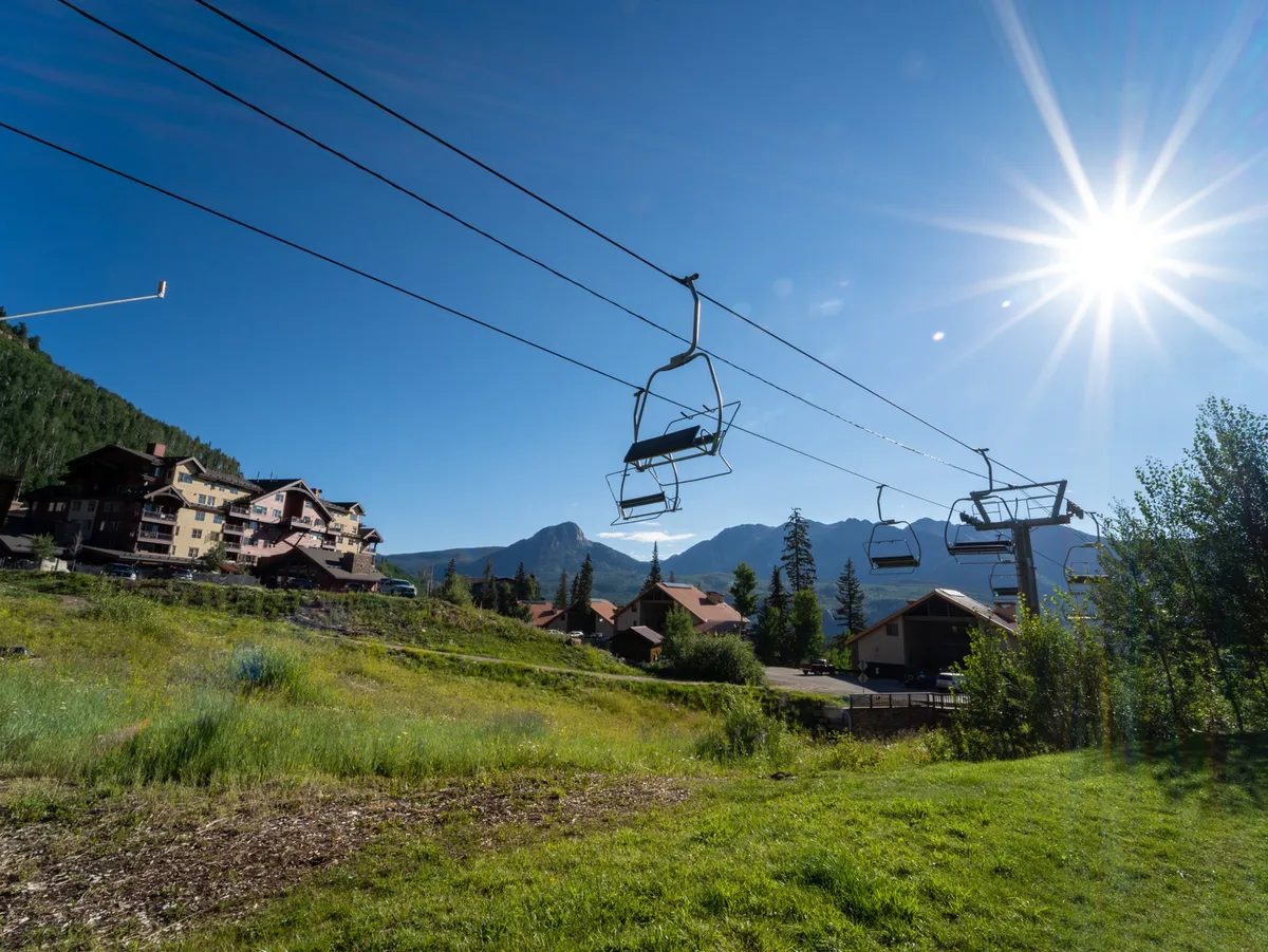 A ski lift glides above lush green grass with a dramatic mountain range in the background, as residential buildings dot the landscape. The scene transitions naturally from winter activities to summer possibilities, like hiking and biking.