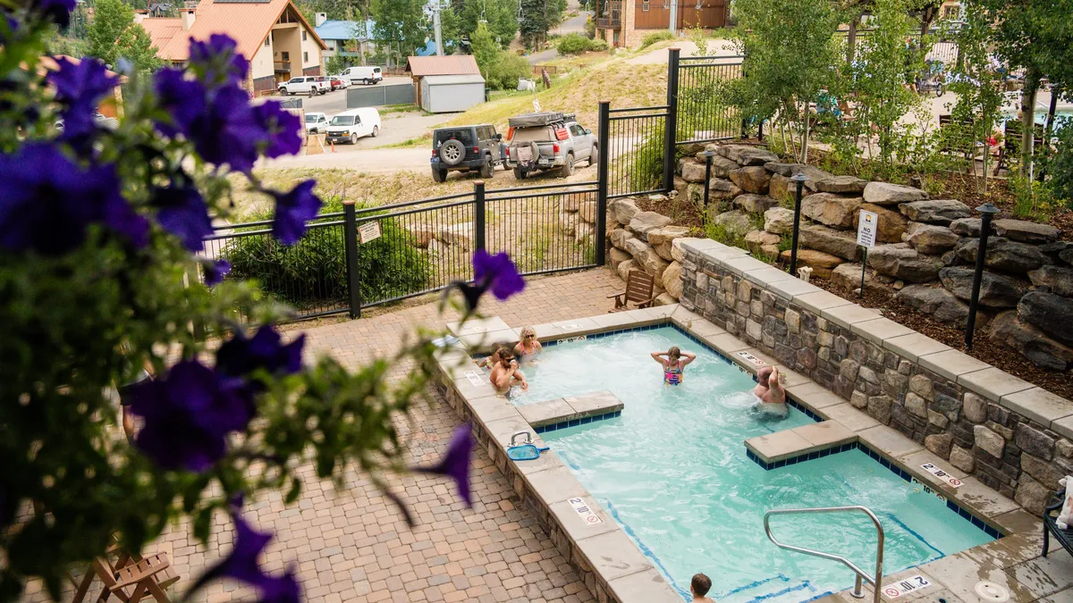 A vibrant outdoor swimming pool surrounded by blooming flowers, with people enjoying the pool and relaxing on a sunny day. The inviting atmosphere hints at socializing and unwinding, with resort amenities evident.