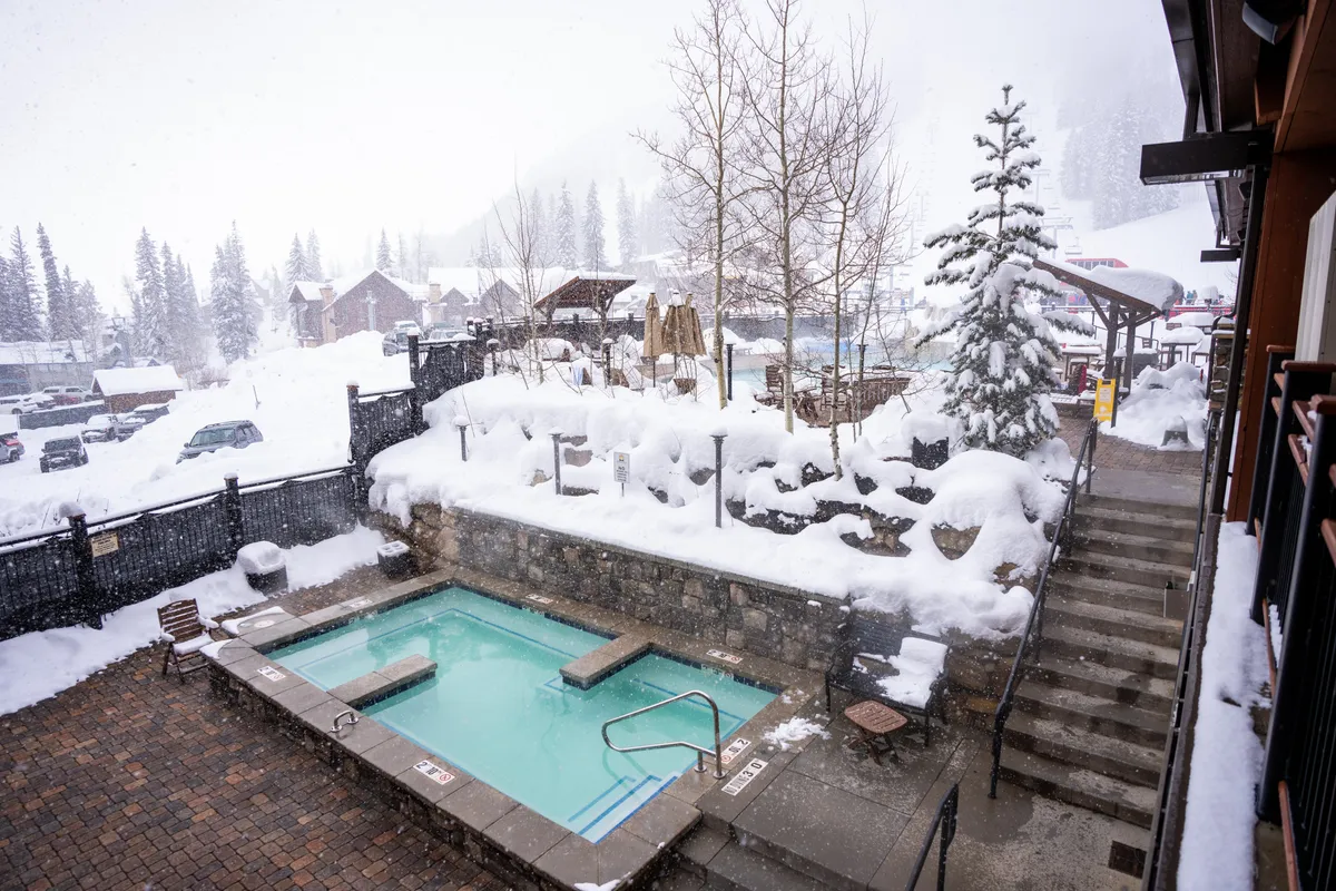 An inviting outdoor hot tub sits amidst a snow-covered landscape and tall trees, creating a serene setting for relaxation. The warm steam contrasts beautifully with the snowy backdrop, suggesting comfort after a day of wintry adventure.