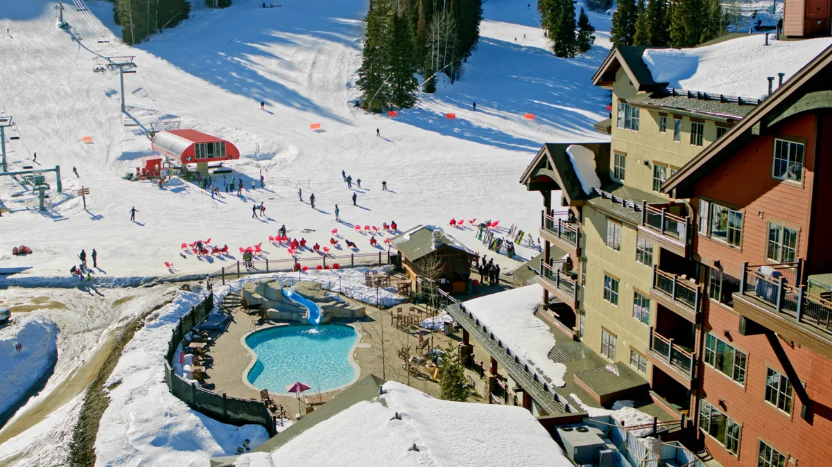 An exciting exterior scene featuring active ski slopes dotted with skiers and snowboarders, a heated outdoor pool with people relaxing, a prominent lodging building in the background, and snow-covered trees that highlight the winter resort atmosphere.