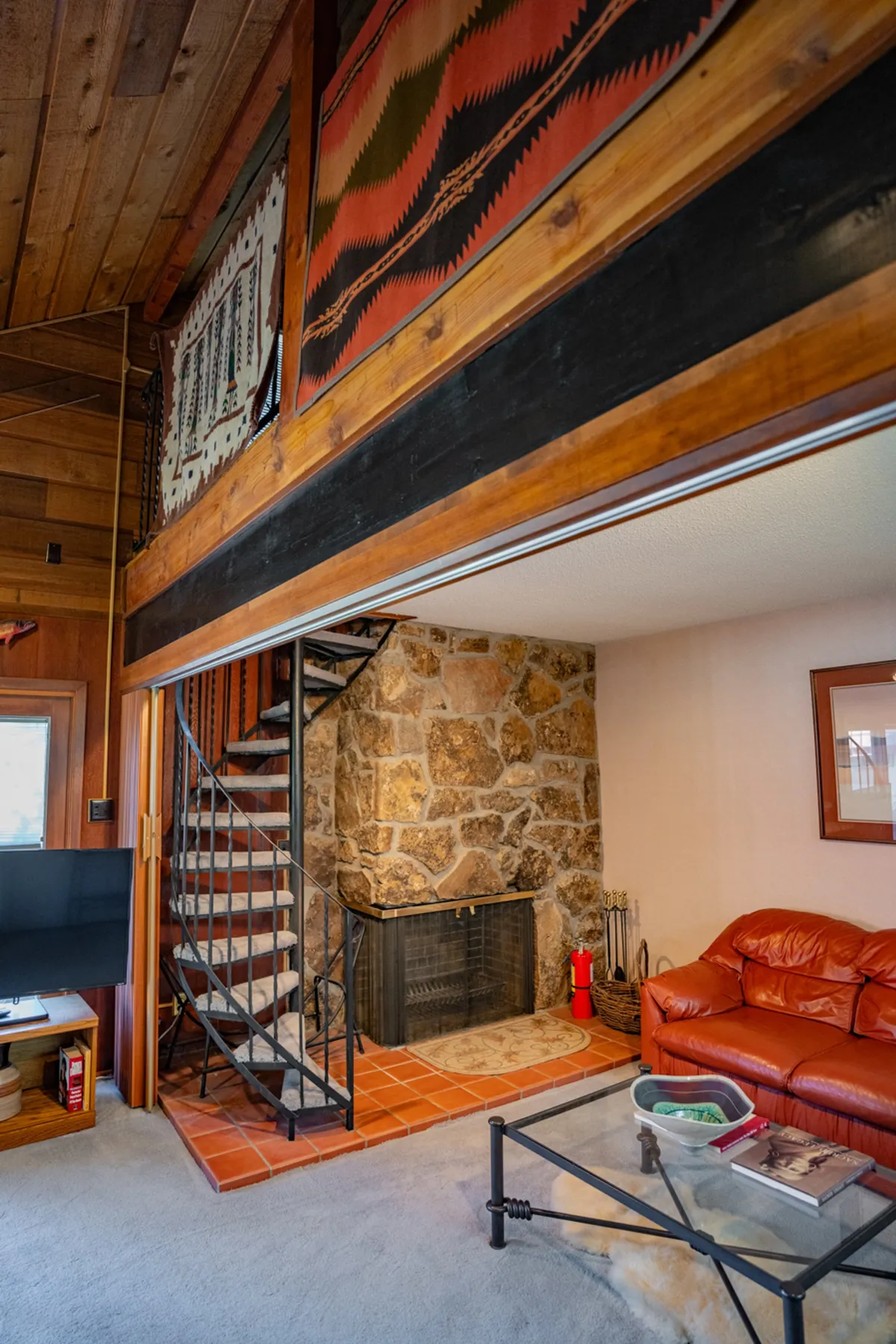This image captures a warm, open-concept living space featuring a prominent stone fireplace, a red leather sofa, a wooden spiral staircase, and woven wall hangings. You can see the living area seamlessly flowing into the dining area, embodying the cozy and communal atmosphere described.