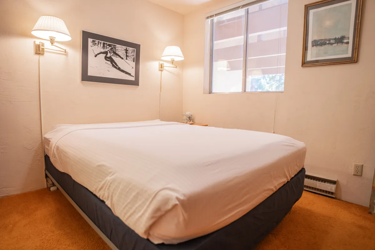 This image features a queen-sized bed with inviting linens, framed artwork adorning the wall, a window streaming in natural light, and wall-mounted lamps. The room exudes a calm and restful mood, ideal for unwinding after an active mountain day.