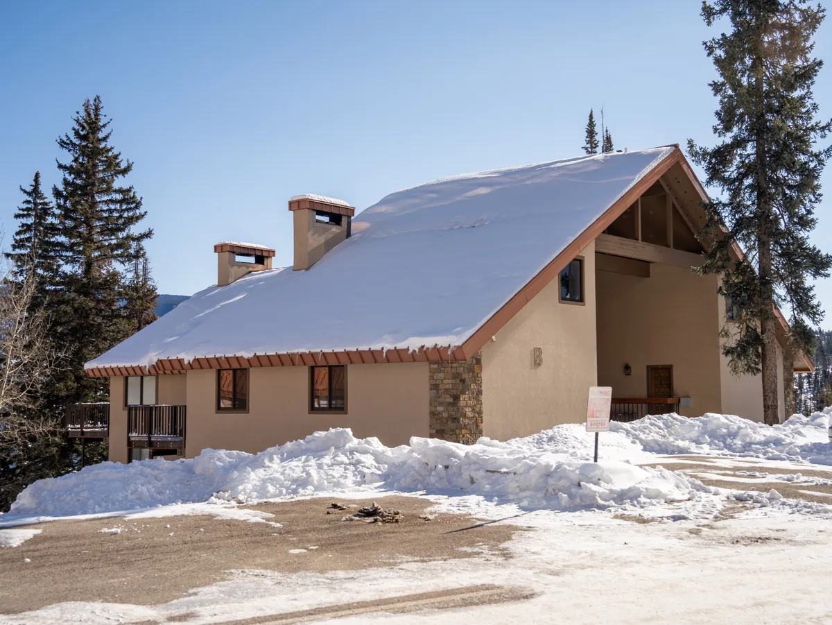 A charming exterior view of the building with a snow-covered roof, stone chimney, and wooden balconies surrounded by tall pine trees. The area is blanketed in fresh snow, evoking the festive and relaxing emotions of a mountain getaway.