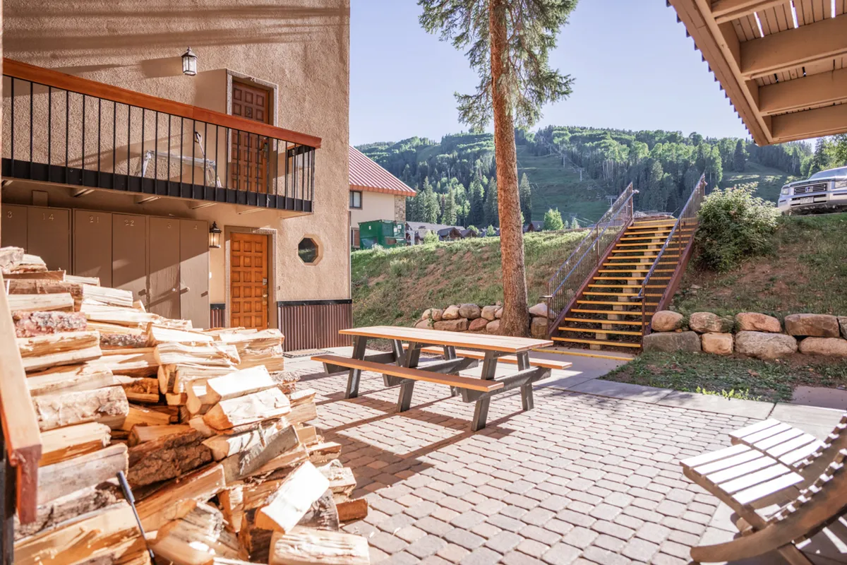 This exterior image features a rustic woodpile, a picnic table, a staircase leading up to the lodging, and a scenic backdrop of the surrounding landscape. The area invites guests to enjoy the outdoors, whether for morning coffee or relaxing après-ski moments.